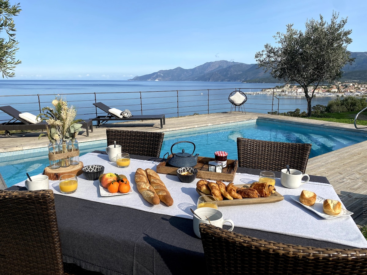 A dining table is set outdoors, featuring a variety of breakfast items including pastries, fruits, and beverages. The table overlooks a tranquil pool, with sun loungers arranged nearby. Scenic views of the coastline and distant mountains are visible, complemented by clear blue skies.
