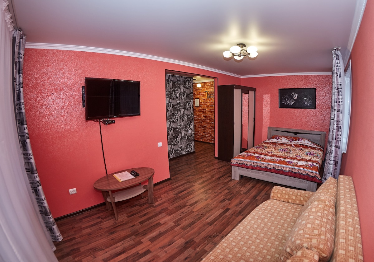 The living area features warm coral walls and a spacious bed with a colorful patterned bedspread. A wall-mounted television is present, along with a cozy couch. Wood flooring complements the room, which is softly illuminated by overhead lighting.