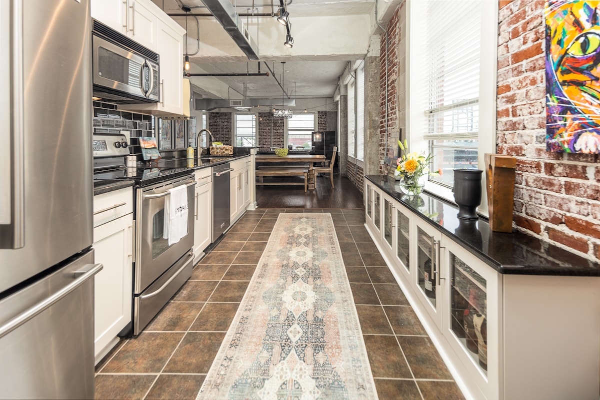 An open kitchen features stainless steel appliances, including a microwave and refrigerator, complemented by granite countertops. A patterned area rug adds warmth to the space, while large windows provide ample natural light, illuminating the brick walls and wooden dining area in the background.