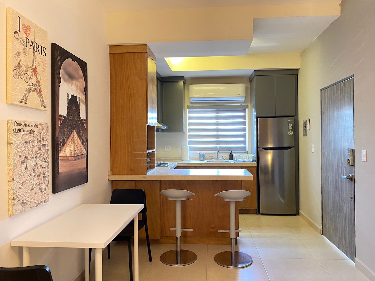 A modern kitchen area is showcased with sleek cabinetry and stainless steel appliances. Two bar stools are positioned at a countertop, while a small dining table is situated nearby. Mounted artwork adds a touch of charm to the neutral-toned walls, enhancing the overall spaciousness.