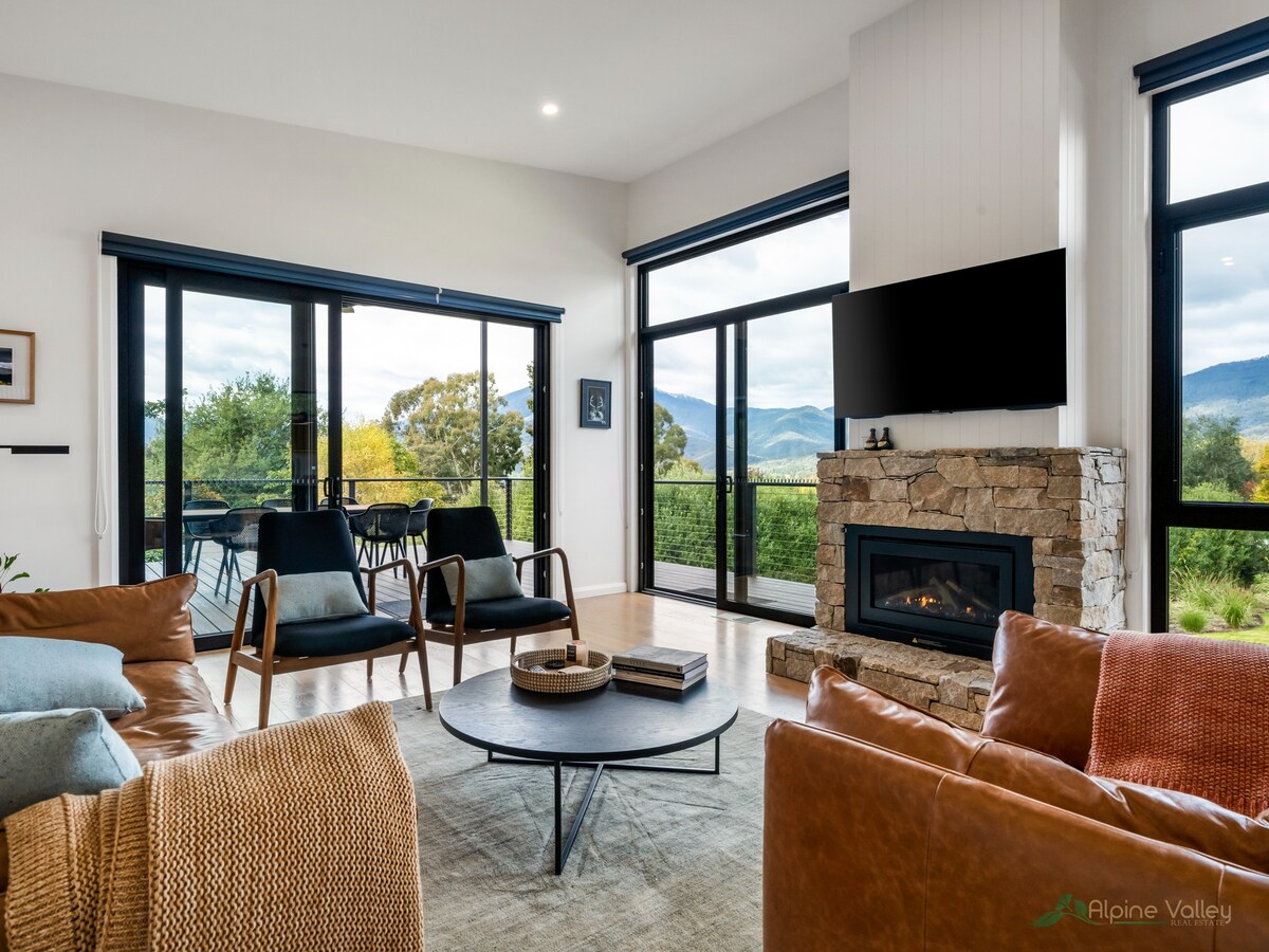 An open living area features large windows that provide mountain views. A stone fireplace is located on one side, while modern seating surrounds a circular coffee table. Warm tones from the furnishings create a comforting atmosphere, with light filling the space.