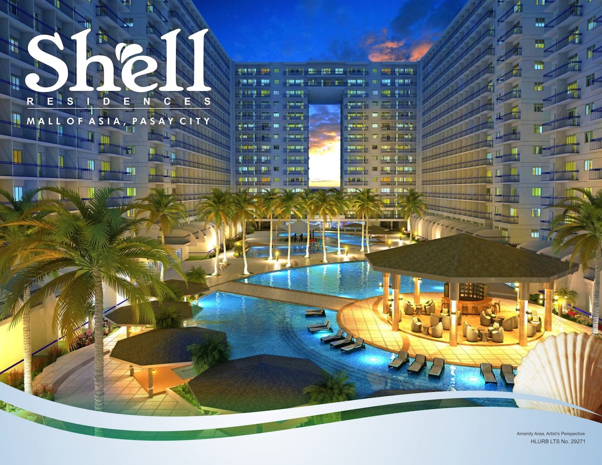 The image depicts the Shell Residences exterior, showcasing multiple building facades surrounding a spacious pool area. Palm trees frame the scene, and a pavilion with seating is visible. The pool features loungers arranged around it, creating an inviting atmosphere for guests.