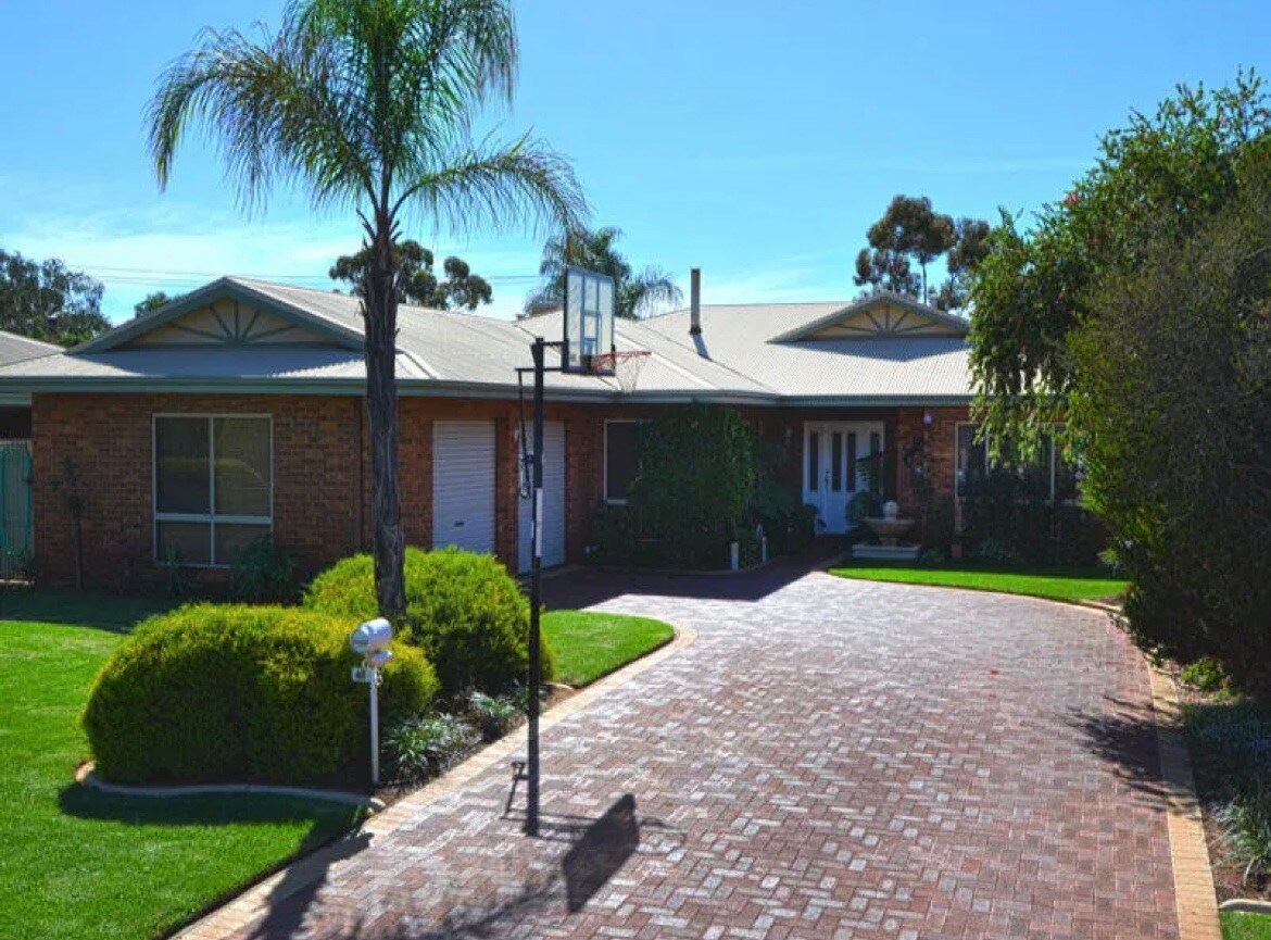 A single-story home is surrounded by well-maintained greenery and features a brick pathway leading to the entrance. A basketball hoop is positioned near the driveway, and tall palm trees provide shade against the clear sky.