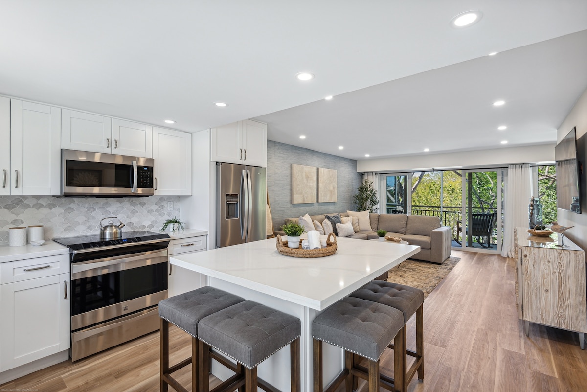 A modern kitchen and living area are featured in an open concept layout. Sleek white cabinets and stainless steel appliances complement a central island with bar seating. Natural light filters in through sliding glass doors, which lead to a balcony surrounded by greenery.