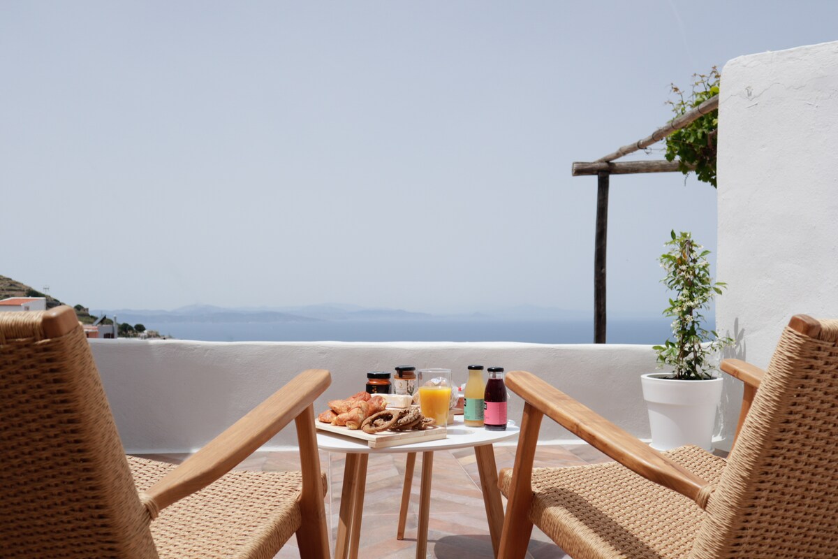 A cozy outdoor seating area is presented, featuring two wicker chairs facing a small table. The table is adorned with an array of breakfast items, including pastries and beverages. A scenic view of the sea and coastline is visible in the background.