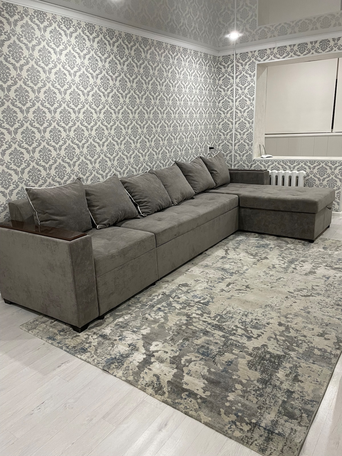 A sectional sofa in a neutral grey tone is arranged on a patterned rug. The wall behind features a classic damask pattern in light grey and white, while soft lighting enhances the space.