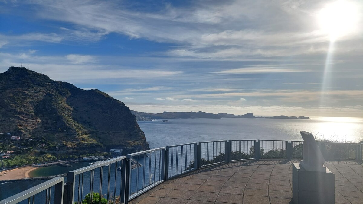 A panoramic view captures the coastline, with mountains rising from the shore and gentle waves lapping at the beach. The sun's rays cast a warm glow over the scene, highlighting the serene waters and distant islands. The observation deck features a railing, offering a safe vantage point.