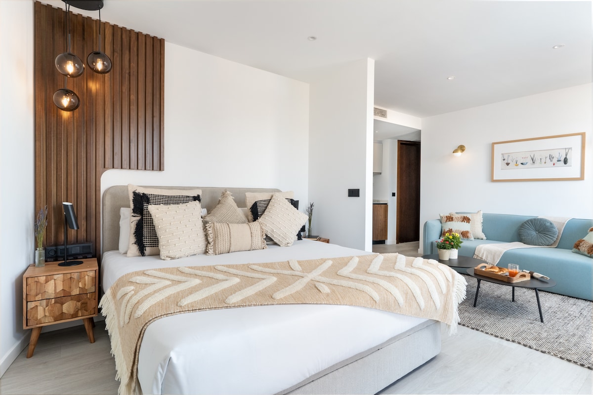 A modern bedroom features a spacious bed adorned with decorative pillows and a textured throw blanket. Stylish wall lighting complements the warm wood accents. A cozy seating area with a light blue sofa and a small table adds to the inviting atmosphere.