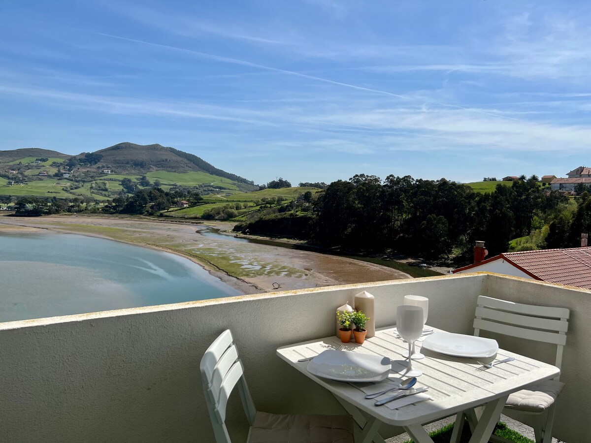 A dining table set for two is positioned on a terrace, offering views of the ría and the surrounding hills. Green fields and trees are visible in the background, with a calm body of water in the foreground under a clear blue sky.