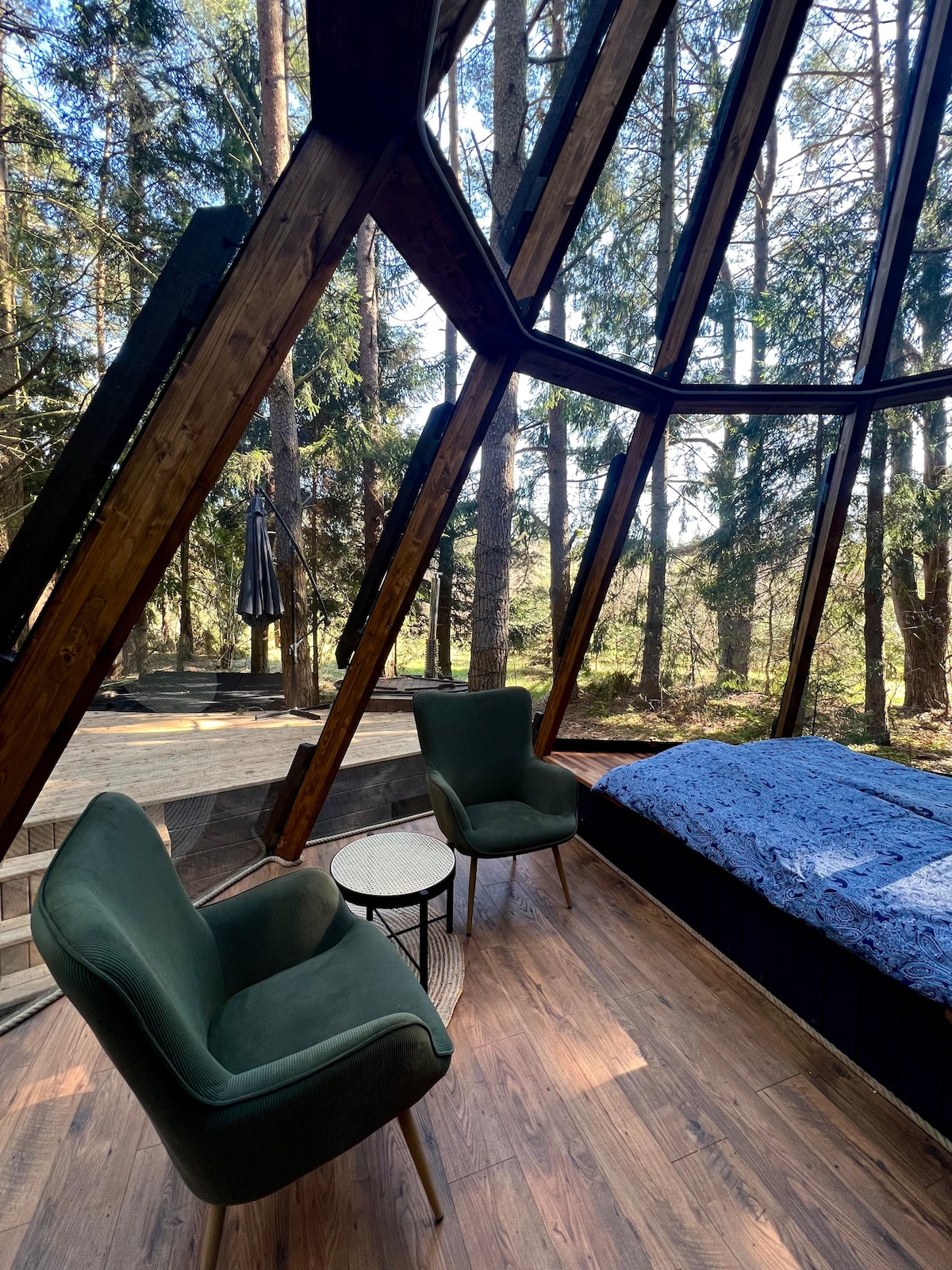 A cozy interior features large glass walls, allowing natural light to fill the space. Two green chairs are positioned beside a small round table, creating a comfortable seating area. A spacious bed with blue bedding is visible, with views of surrounding trees enhancing the tranquil atmosphere.