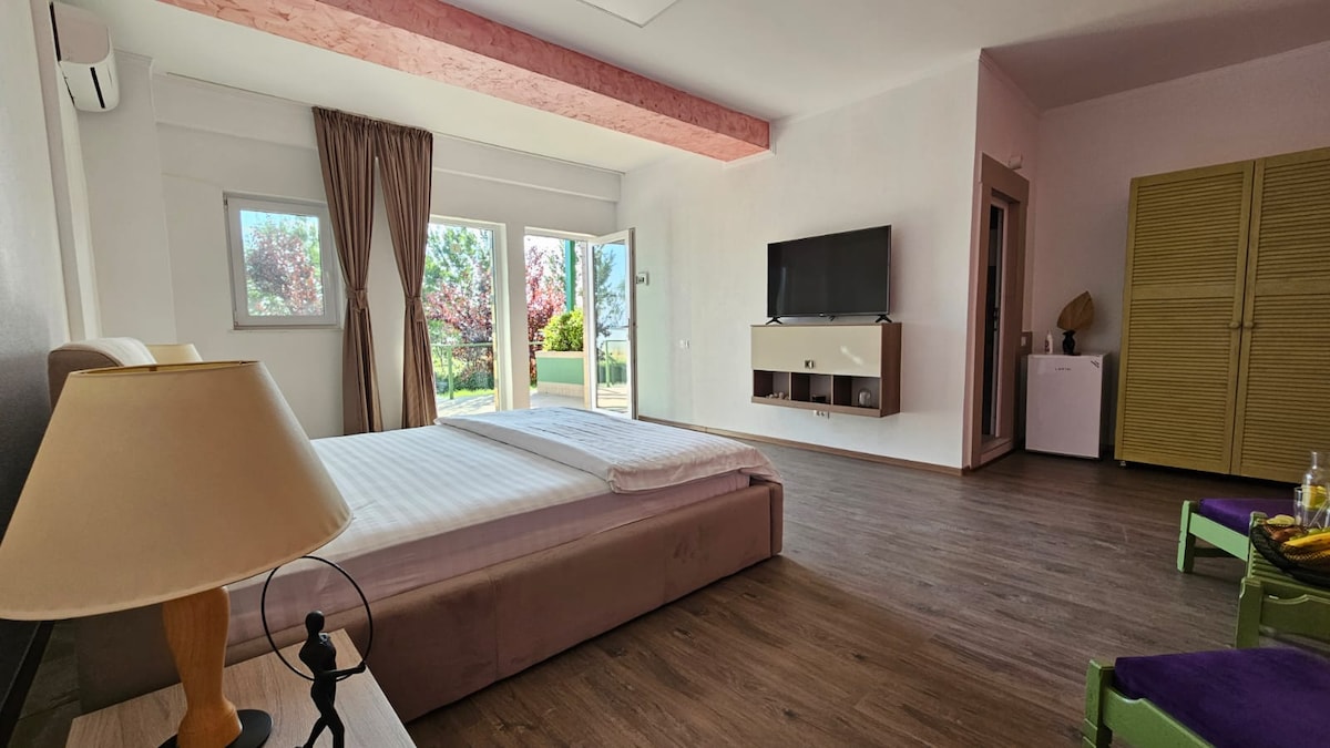 A light-filled room features a spacious bed with neutral linens and a decorative lamp on a bedside table. A flat-screen television is mounted on the wall, while large glass doors open to a garden view. A wardrobe and mini-fridge are visible, enhancing convenience.
