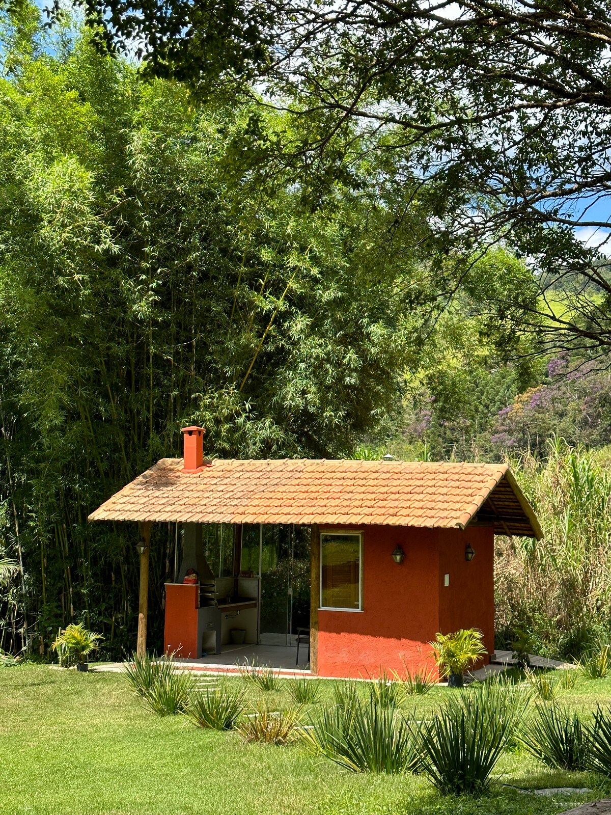 A small, rustic cabin is set amidst lush greenery, featuring a thatched roof and a vibrant orange facade. The structure is complemented by a porch and surrounded by garden areas with native plants, creating a serene and inviting outdoor environment.