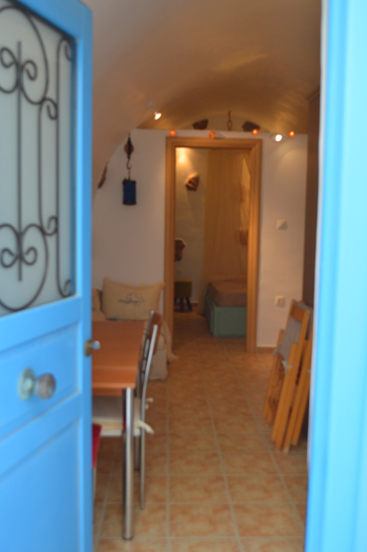 A blue door frames the entrance to the studio, leading into a cozy interior. Natural light illuminates the warm-toned walls and tiled flooring. The dining table is visible on the left, while a serene sleeping area is accessed at the end of the hallway.
