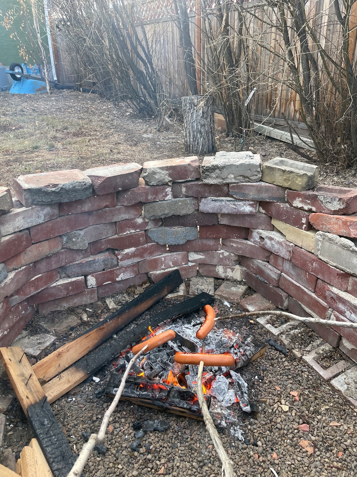 A circular fire pit crafted from stone sits on a gravel surface, with burning logs and hot dogs skewered on sticks. Surrounding vegetation includes bare branches and a wooden fence in the background, suggesting a natural outdoor setting.