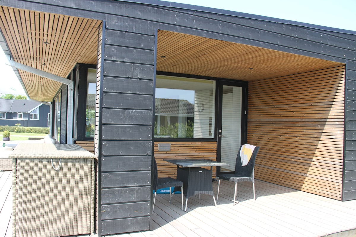 An outdoor seating area is presented, featuring a small table and a single chair. The space is bordered by wooden paneling and large glass windows that allow natural light to fill the area. A woven storage unit is positioned nearby.