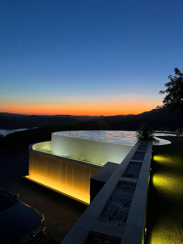 An infinity edge pool is seen gently illuminated by soft lighting, creating a calming ambiance at dusk. The vibrant colors of the sunset reflect off the water, while the surrounding mountains and lake offer a scenic backdrop, enhancing the tranquil evening atmosphere.