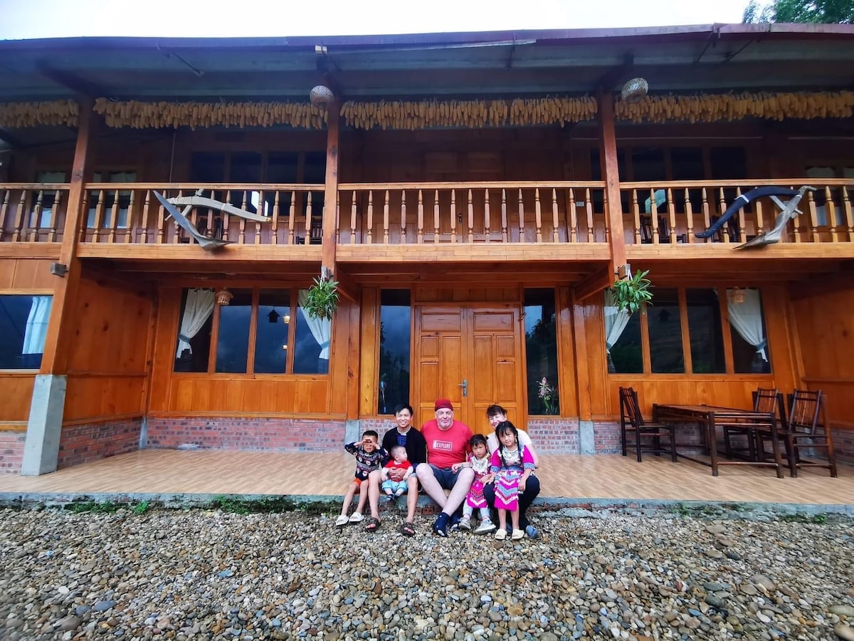 A charming wooden homestay features a spacious front porch with a thatched roof, supported by wooden beams. Families are seated on a gravel surface in front of the house, showcasing a welcoming atmosphere. Plant hangers adorn the porch, adding a touch of greenery.