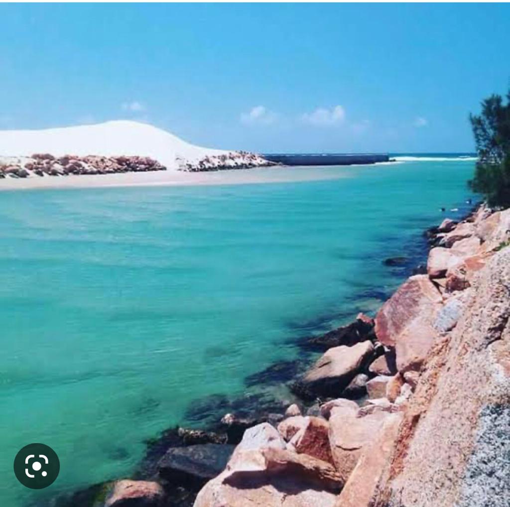 Bright turquoise waters flow beside a rocky shoreline, leading to golden sand dunes in the distance. Clear skies with a few scattered clouds create a serene backdrop for the natural landscape.