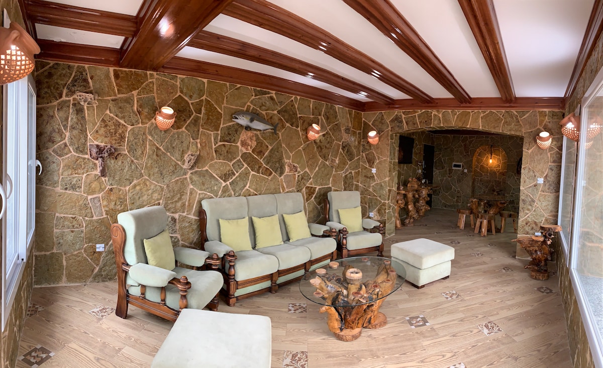 A living area features natural stone walls and wooden beams, accented by soft green seating. A wooden coffee table complements the space, while unique wooden decor adds character. Large windows allow light to fill the room, creating a serene atmosphere for relaxation.