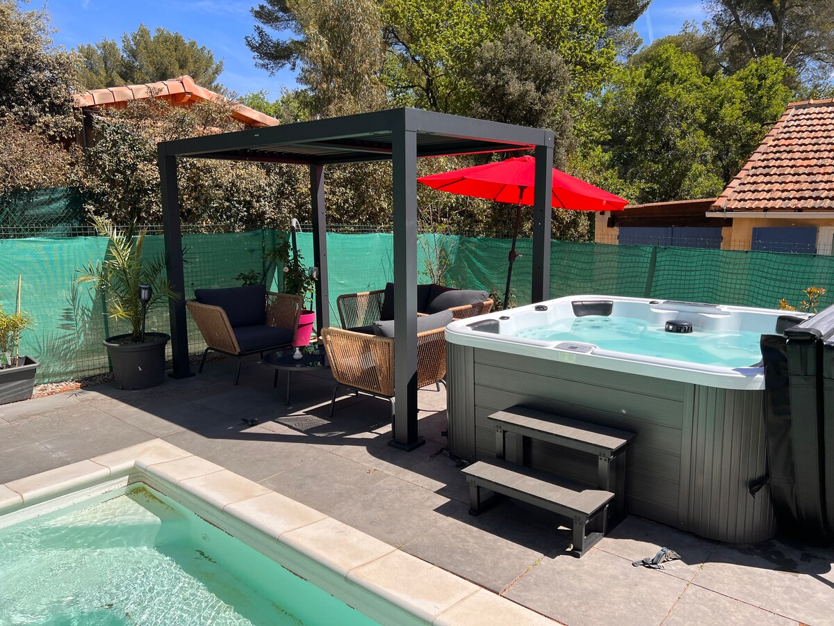 A shaded seating area is provided near a jacuzzi, with comfortable chairs positioned under a black gazebo. An umbrella offers protection from the sun. The jacuzzi, surrounded by paving stones, invites relaxation amidst greenery and a fenced backdrop.