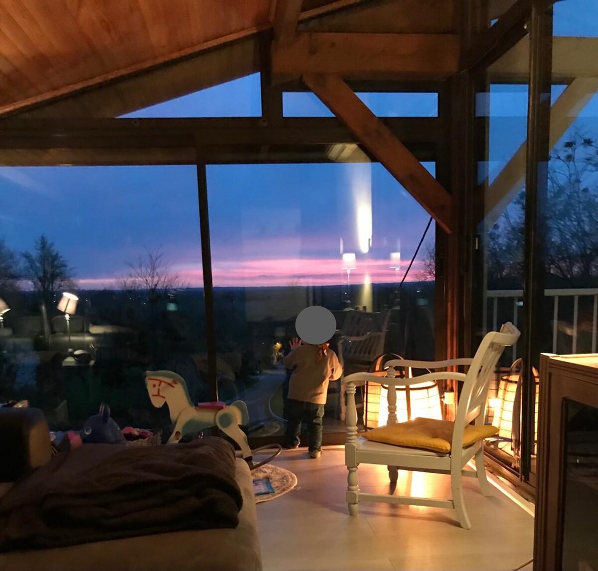Large windows with wooden beams frame a child gazing at a colorful sunset. Soft lighting accents the cozy seating area, which includes a white rocking chair and a plush rug. Views of the surrounding landscape create a serene atmosphere inside the home.