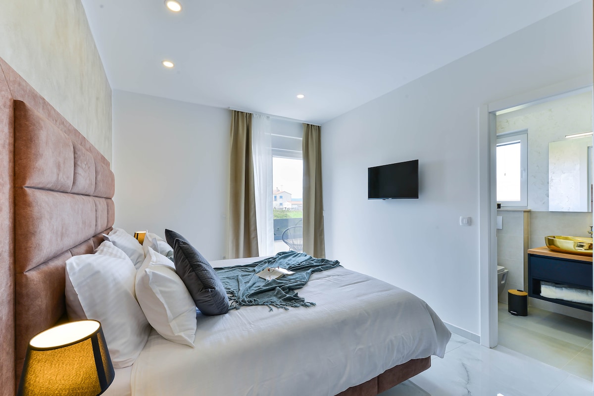 A contemporary bedroom showcases a spacious bed with a muted headboard and neatly arranged pillows. Soft light fills the room through a large window dressed with light curtains. A flat-screen television is mounted on the wall, and an en-suite bathroom is partially visible through an open door.