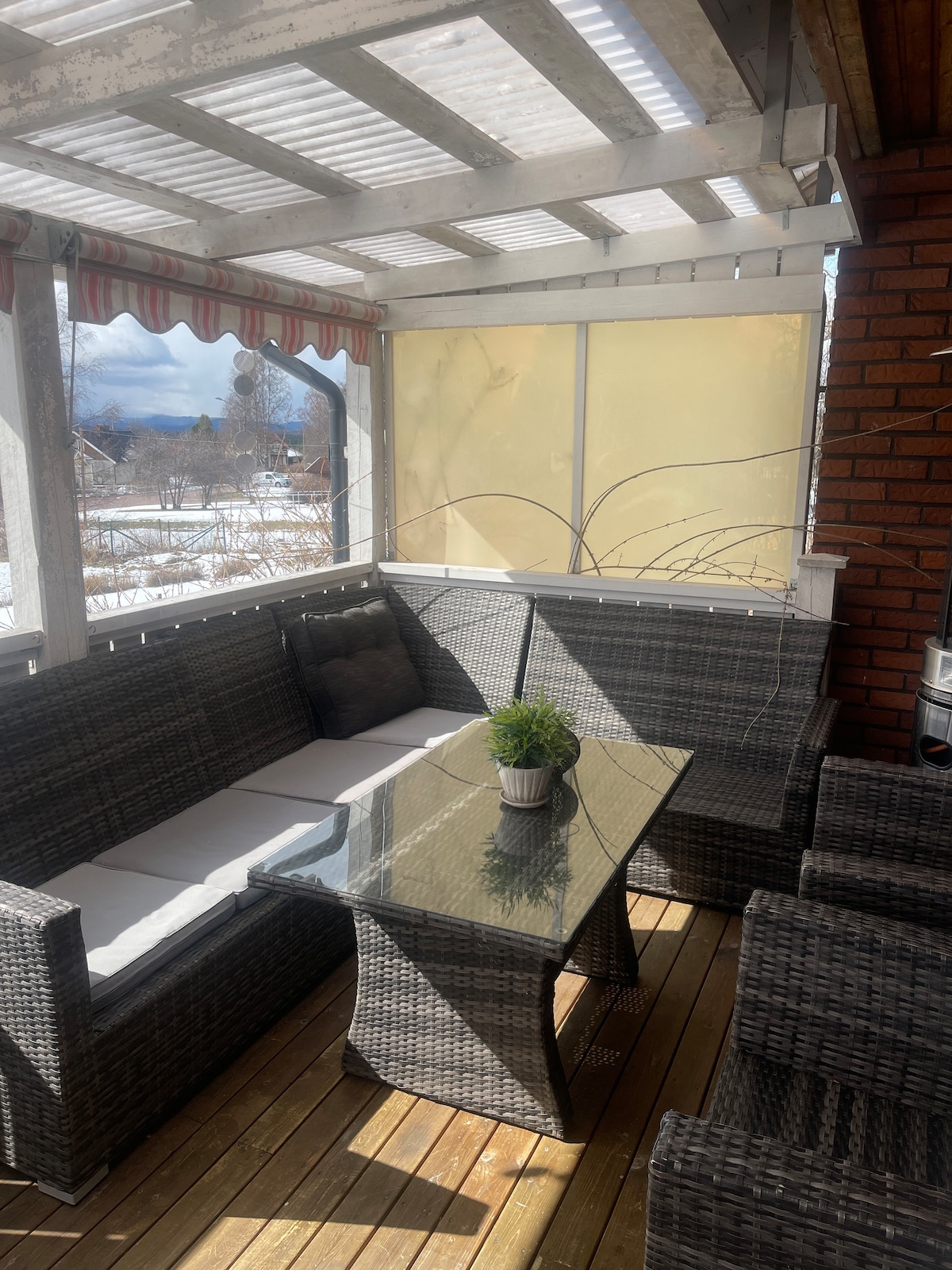 A covered seating area features a woven sofa with light cushions surrounding a glass-top table, creating a cozy space for gatherings. Natural light filters through the partially transparent window coverings, highlighting the wooden floor and offering a view of the outdoors.