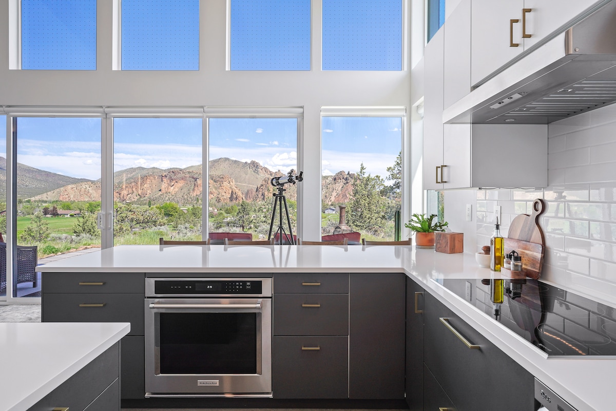 A modern kitchen features sleek countertops and dark cabinetry, complemented by large windows that frame expansive views of Smith Rock. Natural light floods the space, highlighting a variety of kitchen appliances and a telescope positioned by the window.