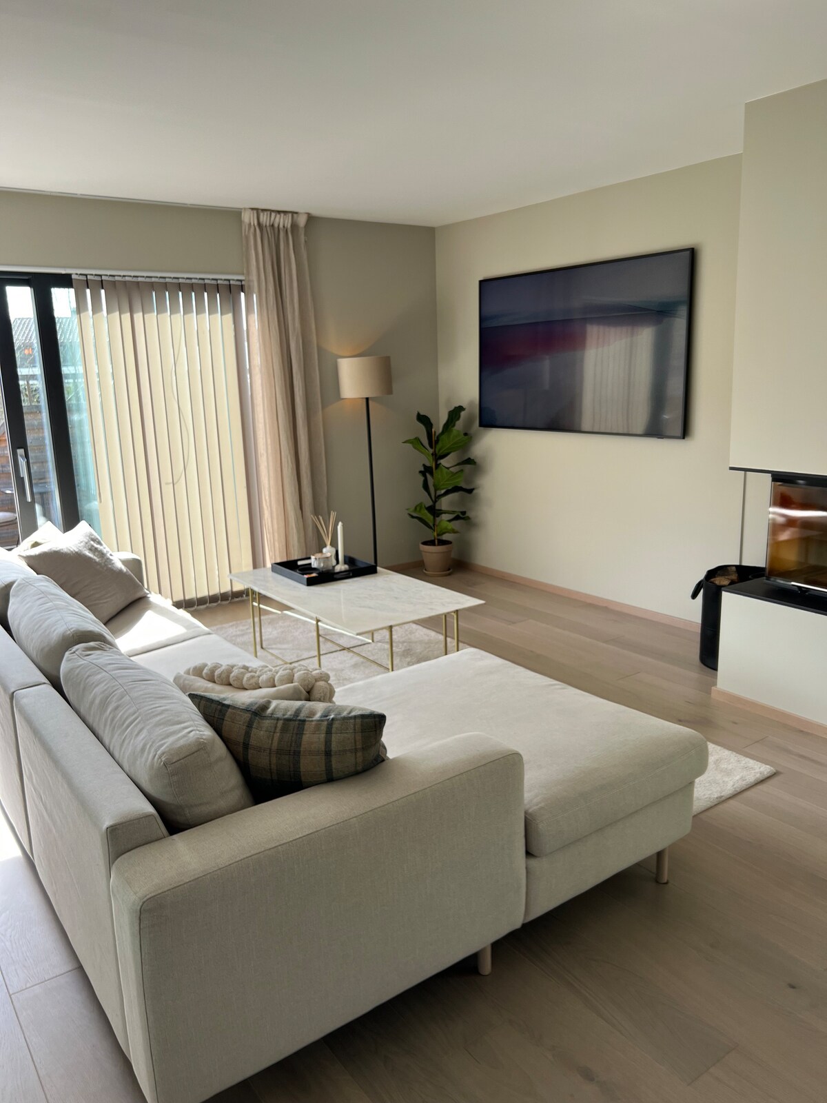A modern living area features a light-colored sofa with decorative pillows, positioned near a sleek coffee table. Floor-to-ceiling windows allow natural light to fill the space, and a green potted plant adds a touch of nature. A mounted flat-screen television is visible on the wall.