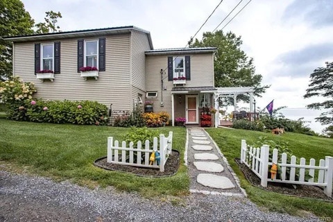 A welcoming entrance to a newly renovated home is framed by a well-maintained yard featuring colorful flower beds and a path made of stepping stones. A white picket fence defines the garden area, enhancing the serene outdoor ambiance.