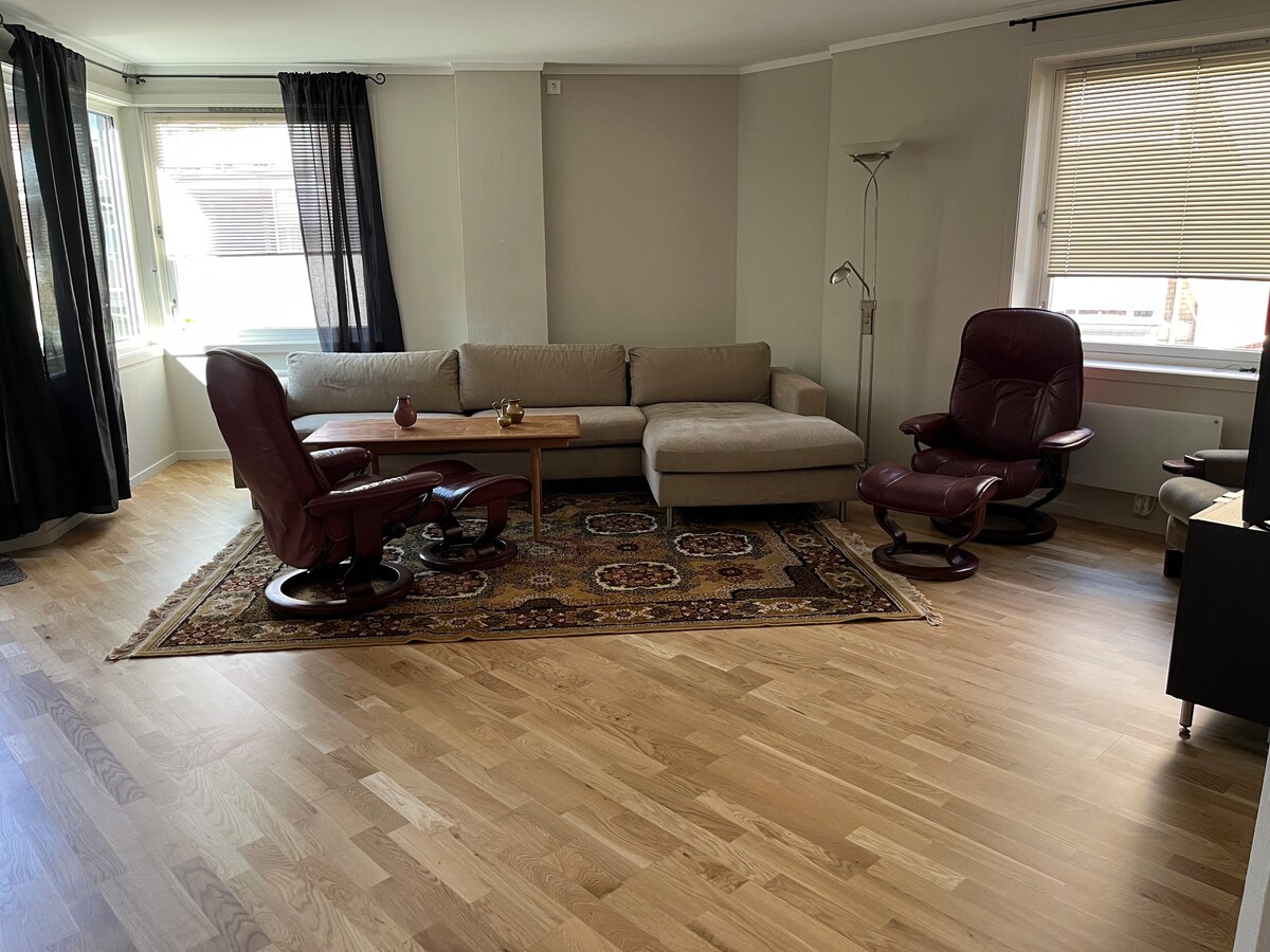 A comfortable living area is presented with a light-colored sectional sofa and two burgundy recliners, arranged around a wooden coffee table. A patterned area rug adds texture to the space, while large windows provide natural light, complemented by dark curtains.