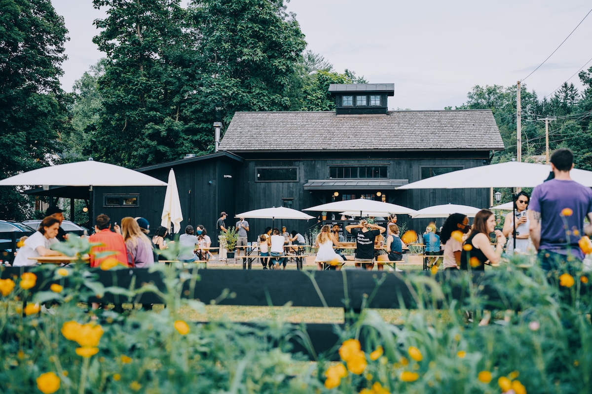 A modern black structure serves as the backdrop for a vibrant outdoor dining area. Guests are seated at tables under large umbrellas, surrounded by flowers and greenery. The atmosphere appears lively, with people enjoying their time in the fresh air.