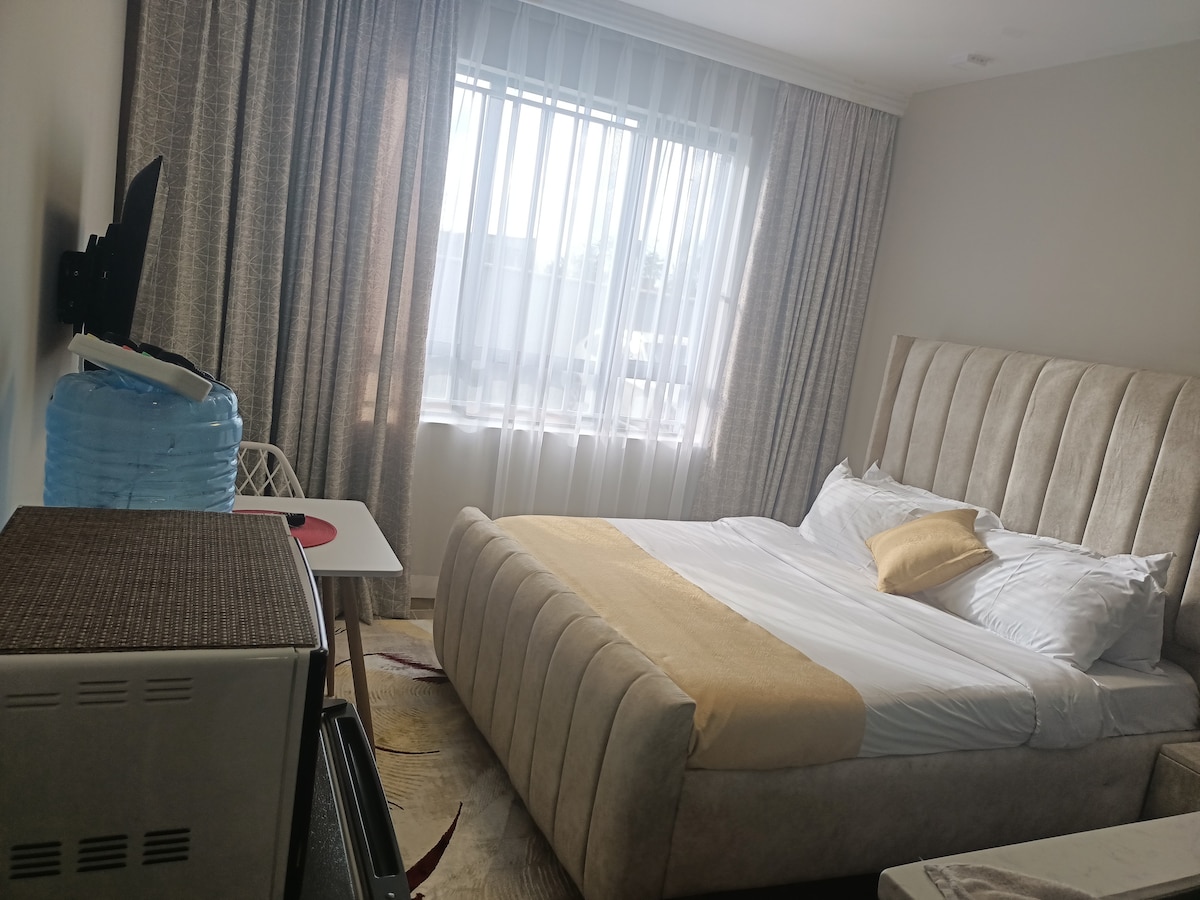 A well-lit bedroom features a spacious bed with a light cream headboard and white bedding. Soft curtains gently filter natural light from a large window. A small dining table is positioned nearby, complemented by a textured rug and a television mounted on the wall.