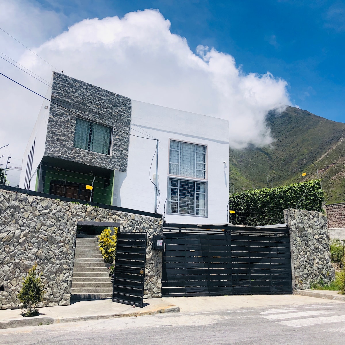 A modern two-story house features a combination of white and stone exterior elements, with large windows allowing natural light. A lush green mountain rises in the background, and a sleek, black gate stands at the entrance, surrounded by manicured landscaping.