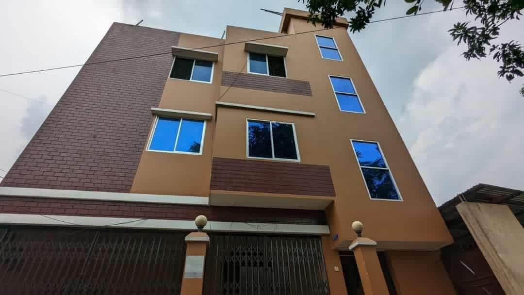 The exterior of a three-story building is displayed, featuring a combination of brown brick and light tan siding. Large windows are present on each floor, reflecting the sky. A decorative fence is positioned at the front, enhancing the building's entrance.