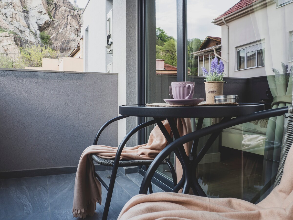 A balcony area is shown with a small black table and two wicker chairs. A soft, beige throw drapes over one chair. A lavender plant in a pot adds a touch of greenery, while a pink cup rests on the table, creating a serene outdoor setting.