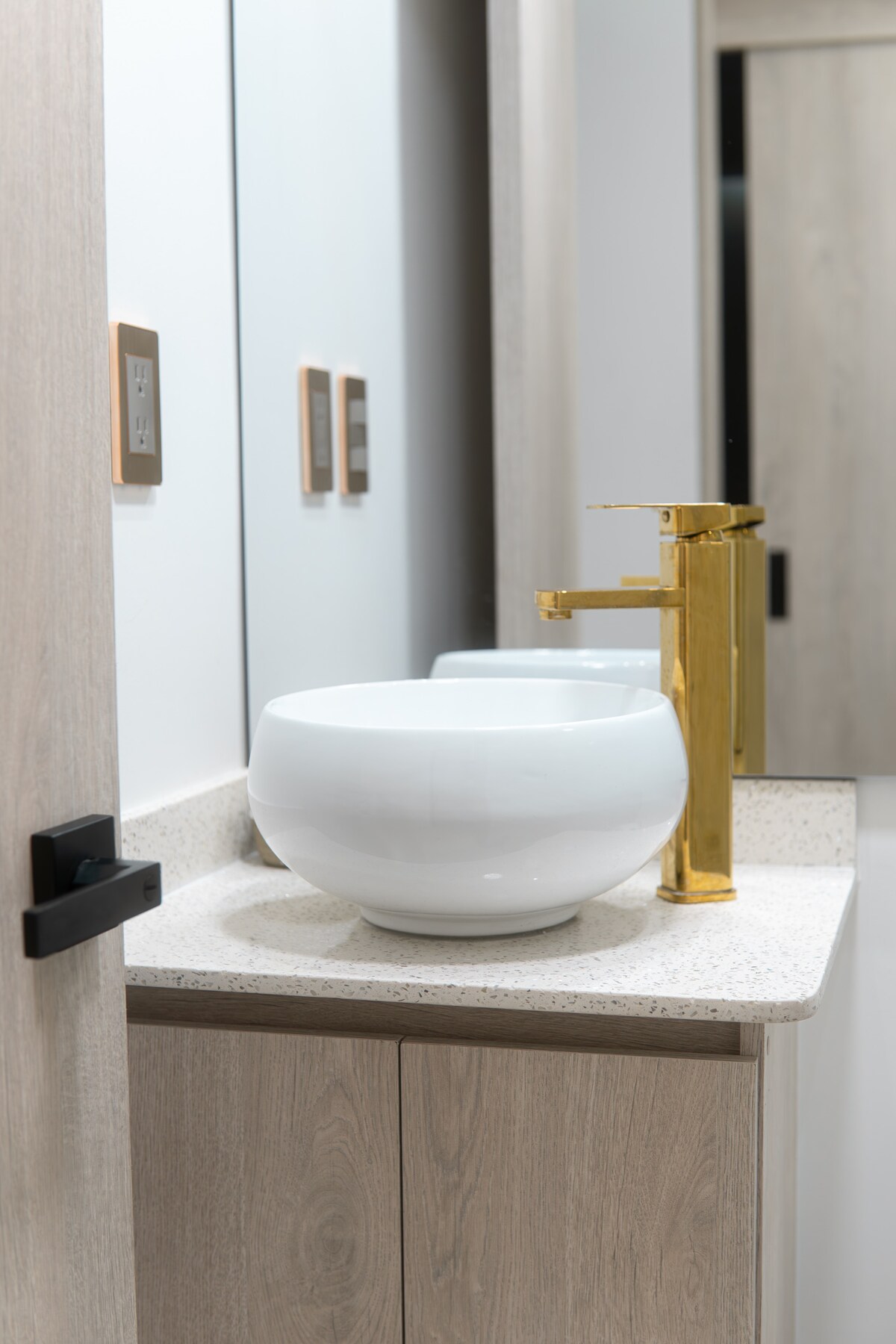 A modern bathroom is presented, featuring a sleek countertop with a white bowl sink. A stylish gold faucet complements the sink, while light wood cabinetry offers additional storage. The mirror reflects natural light, contributing to a bright and airy environment.