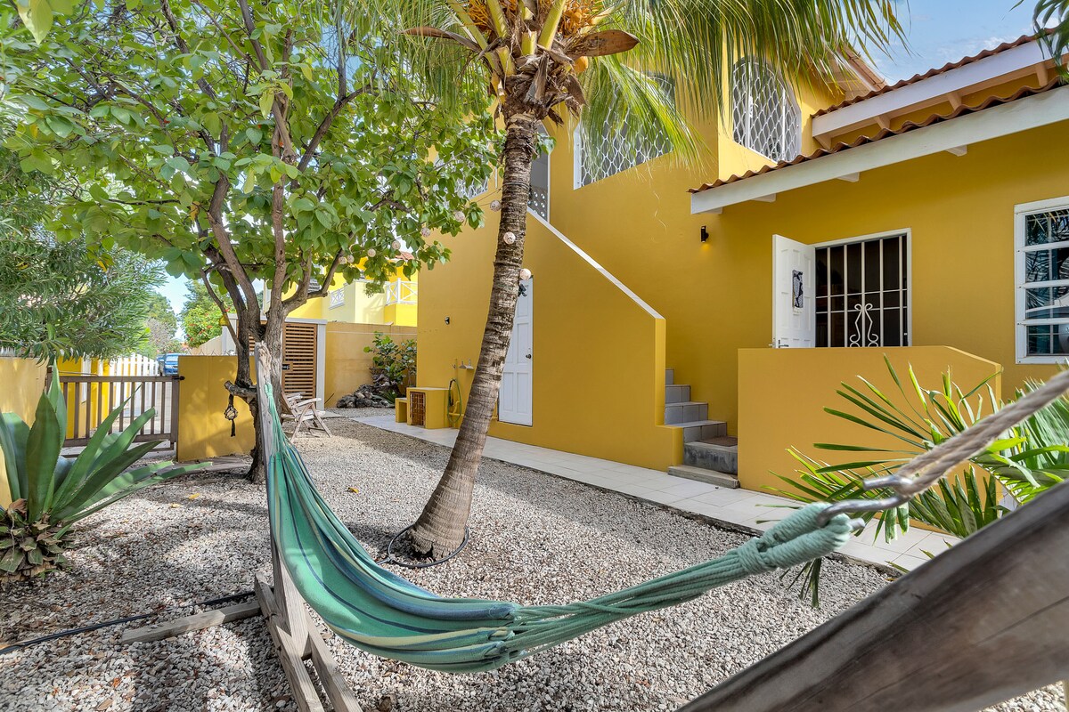 A vibrant yellow exterior is framed by lush greenery and palm trees. A hammock suspended between a tree and a wooden stand invites relaxation in the semi-private garden area. Steps lead to a bright entryway, enhancing the welcoming atmosphere of the studio.