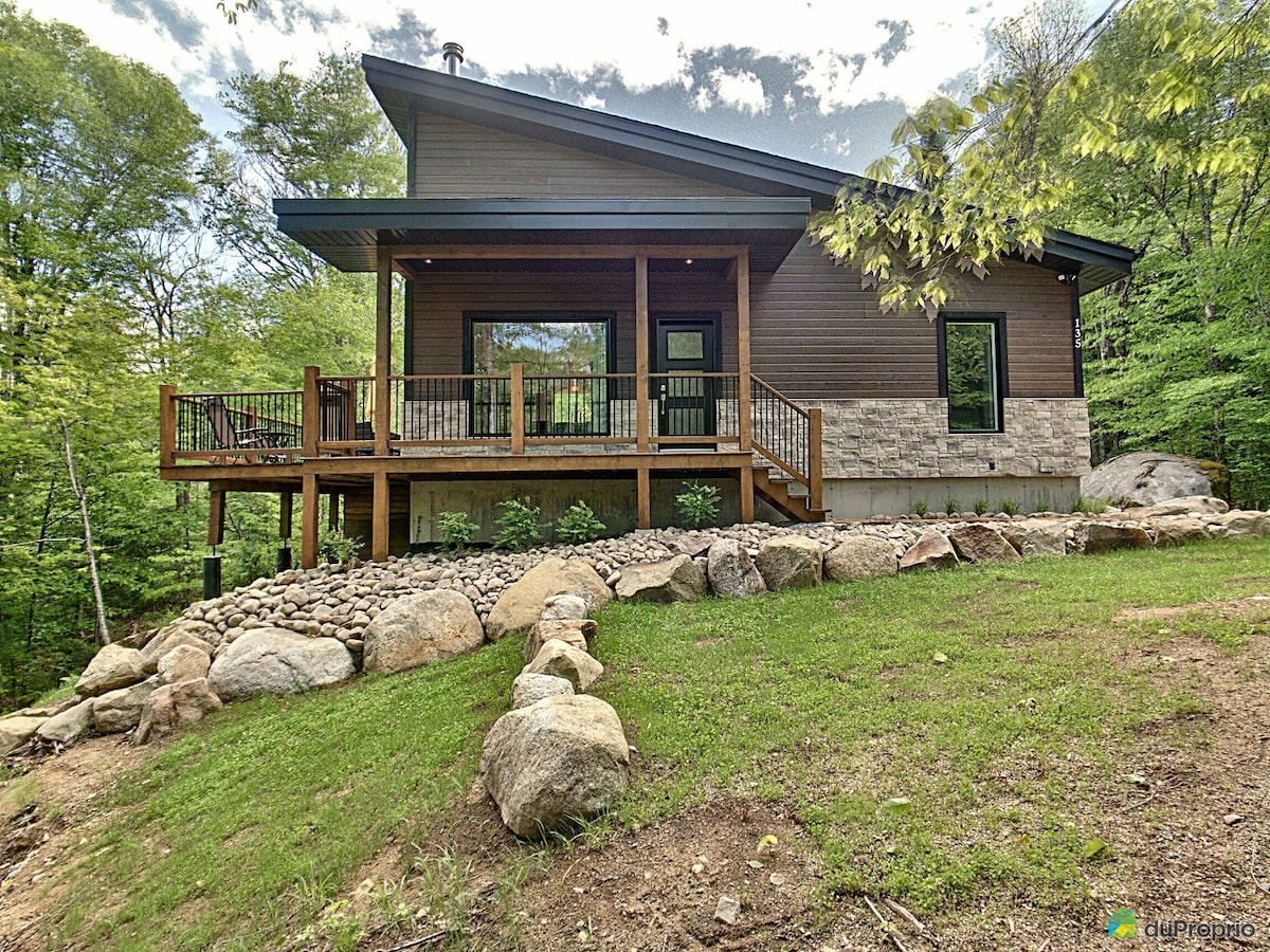 The chalet's exterior is showcased, featuring a blend of stone and wood materials. A spacious deck with railings extends from the front, surrounded by landscaped rocks and greenery. Natural light filters through large windows, enhancing the inviting ambiance of the home.