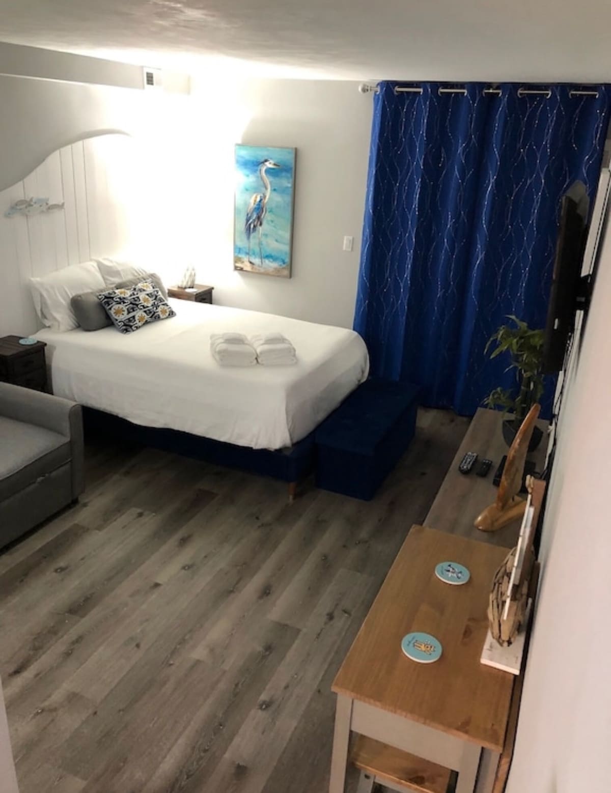 A cozy sleeping area features a spacious bed with white linens and neatly arranged towels. A bright painting of a heron adorns the wall. A comfortable loveseat sits nearby, and blue curtains frame a window, providing a calming contrast to the light wood flooring.
