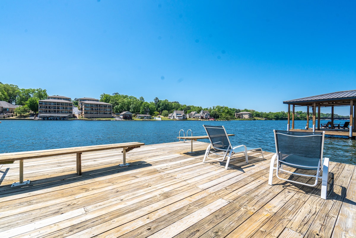 A spacious wooden dock extends over the water, featuring several lounge chairs positioned for relaxation. The calm lake reflects clear blue skies, with green trees lining the opposite shore and several buildings visible in the background.