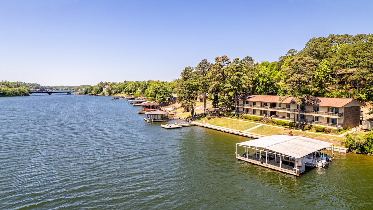 A scenic view of Lake Hamilton is captured, showcasing a peaceful shoreline lined with trees. Several boat docks are visible along the coast, while a quaint condo building stands nearby, surrounded by a spacious lawn that extends to the water's edge.