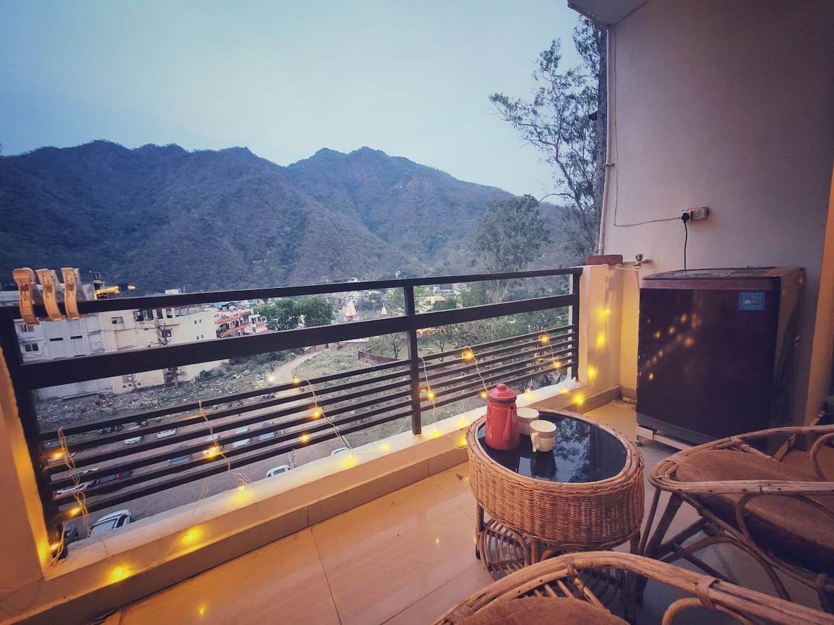 A serene balcony offers a view of surrounding mountains, showcasing a tranquil atmosphere. String lights create a warm glow, illuminating a small table and two wicker chairs. A dark cabinet is positioned against the wall, enhancing the outdoor space for relaxation.