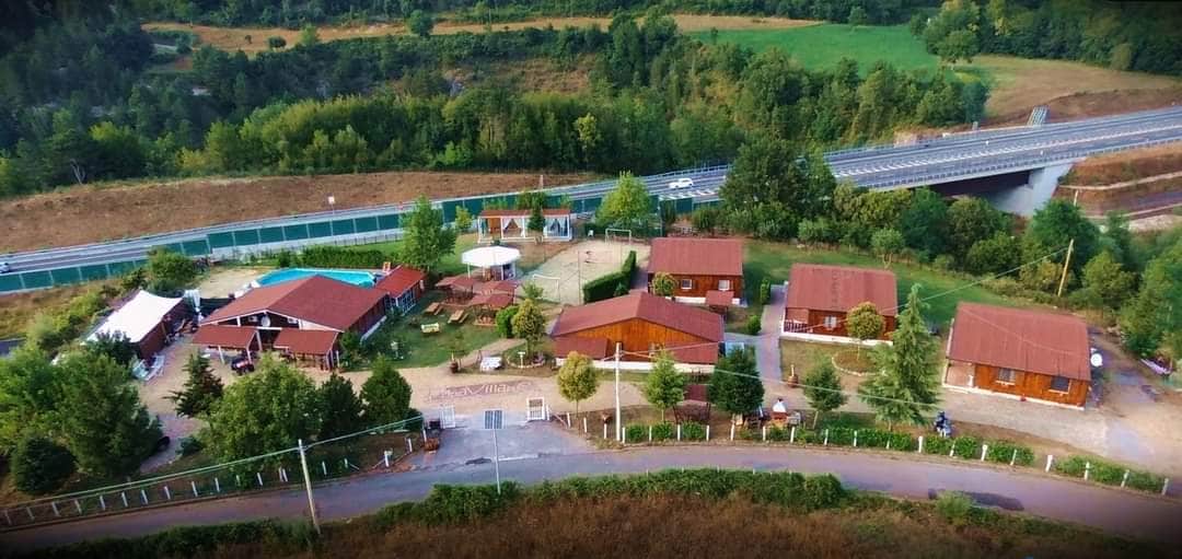 An aerial view captures multiple wooden cabins nestled among green trees, surrounded by open space. A swimming pool can be seen, along with seating areas and sun umbrellas. Nearby, a road is visible, framed by natural landscaping.