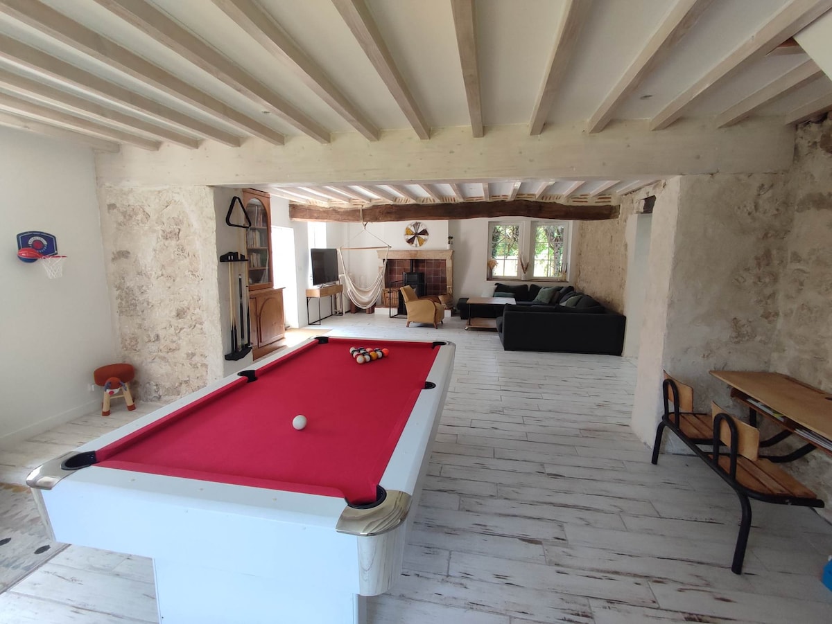 A spacious living area features a bright red billiard table at the center. A comfortable seating arrangement with dark couches is visible in the background, along with a large TV. Natural light enters through multiple windows, illuminating the rustic stone walls and wooden beams.