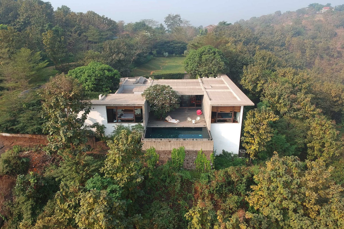 The residence is set amidst greenery, showcasing well-maintained trees and shrubs. An infinity pool extends from the building, reflecting the surrounding landscape. The modern structure features a flat roof and large windows, providing a seamless connection between indoor and outdoor spaces.