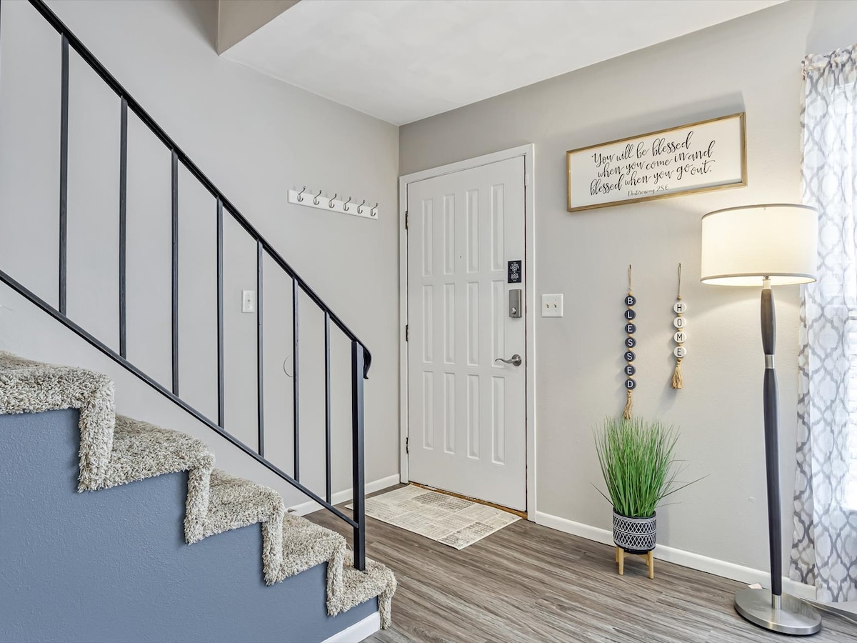 The entryway features a staircase with a textured carpet, leading to the upper level. A front door is positioned on the right, accompanied by a floor lamp and decorative wall hangings. Hooks for coats are mounted on the wall, and a potted plant adds a touch of greenery.