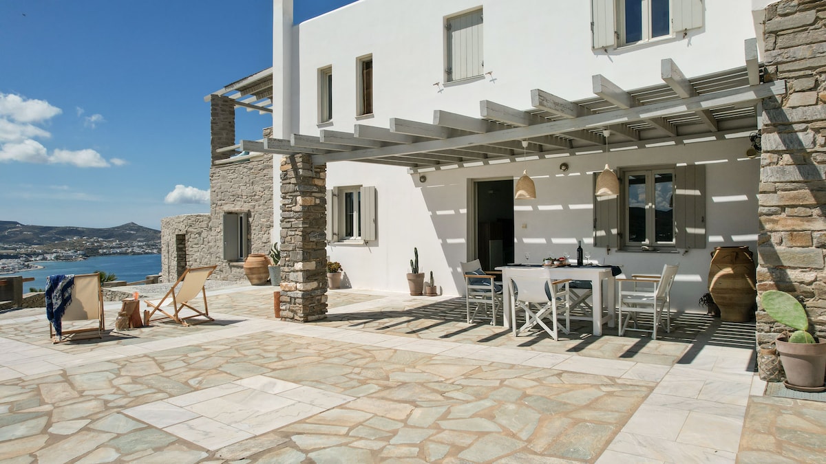 An outdoor terrace is revealed, featuring a spacious dining table set for six. Sunbeds are arranged nearby, with a backdrop of stone walls and large windows. The view overlooks the azure Aegean Sea and the landscape of Parikia, complemented by dappled sunlight through the overhead pergola.