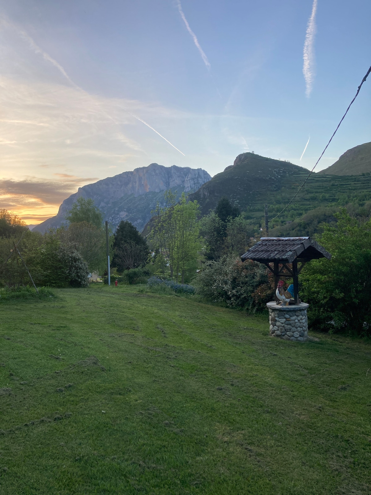 A spacious green yard is framed by lush trees and plants, with a stone well positioned in the foreground. Majestic mountains rise in the background, under a colorful sky that transitions from blue to hints of orange as the sun sets.