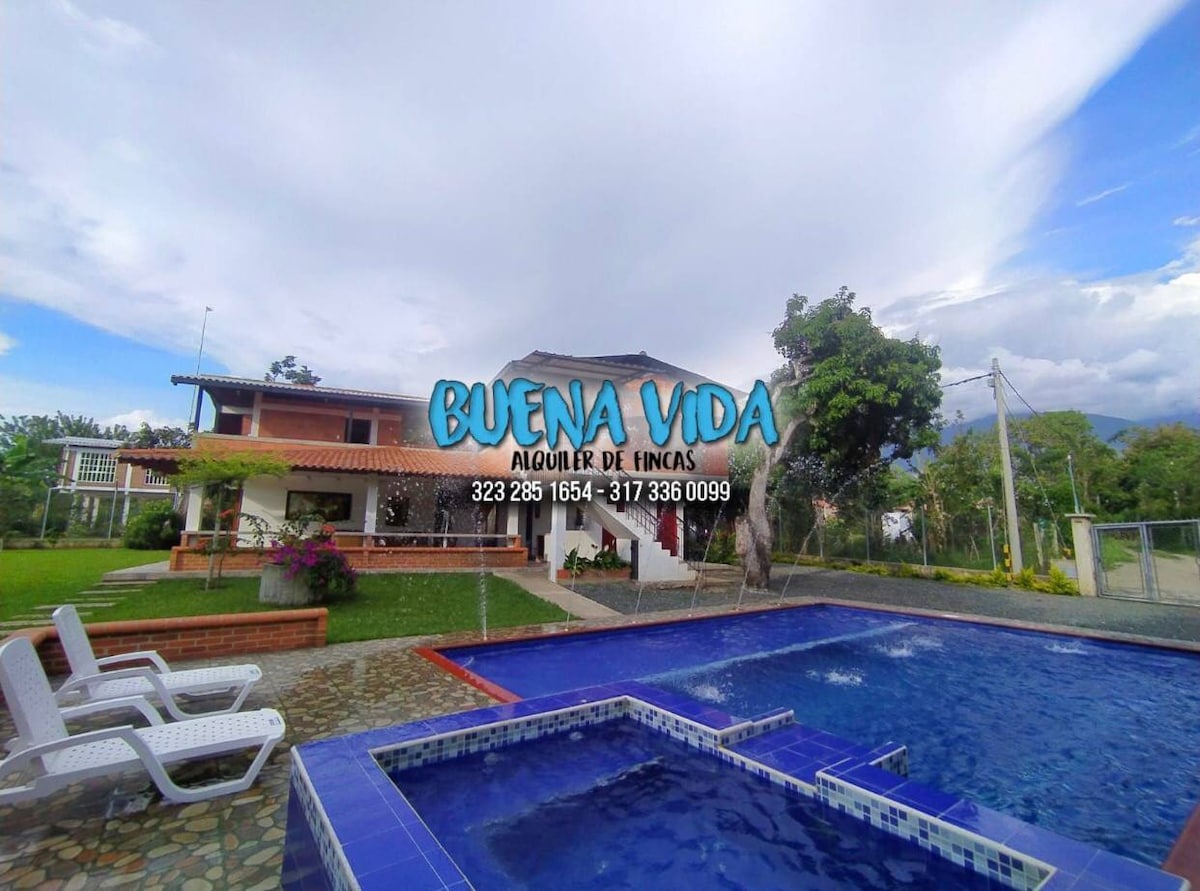 A spacious outdoor area features a swimming pool with a tiled border. White lounge chairs are arranged nearby on a stone patio. The backdrop includes a two-story house with a tiled roof surrounded by greenery and trees, creating a relaxed environment.